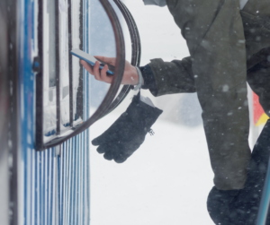 A skier in light snow flurries hold a phone with his bare right hand up to an automated lift ticketing window. His glove hangs freely from his wrist. 